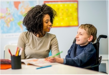 Young boy in a wheelchair writes at a desk as his teacher sits beside him engaging in conversation