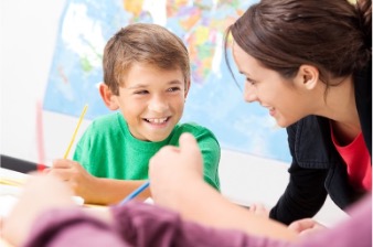 Female teacher leaning over a desk smiling and interacting with a young male student.