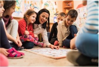 A teacher sitting on the floor pointing to a display board in front of her. Children sit all around her in a circle looking at the board.