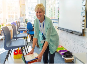 School staff member with a blue lanyard smiles as she puts a stacks of folders out onto desks