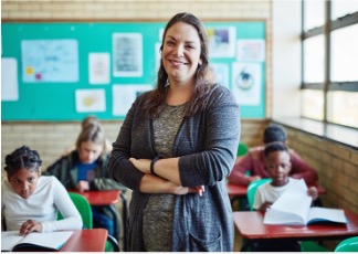 Teacher standing in her classroom smiling at the camera while students behind her work at their desks