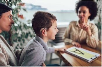 A young caucasian boy and his mom sit across the table from a black woman who is signing. They are all looking at a piece of paper with the letter "A" printed on it which is on the table between them.